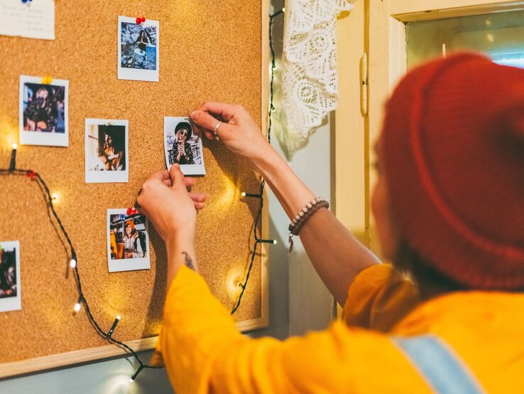 Junge Frau mit roter Mütze und gelbem Pullover pinnt Polaroid-Foto an eine Pinnwand aus Kork mit Lichterkette.