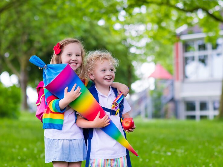 Zwei fröhliche Kinder, eines mit Locken und eines mit Haarspange, halten eine bunte Schultüte im Grünen vor einem Schulgebäude.