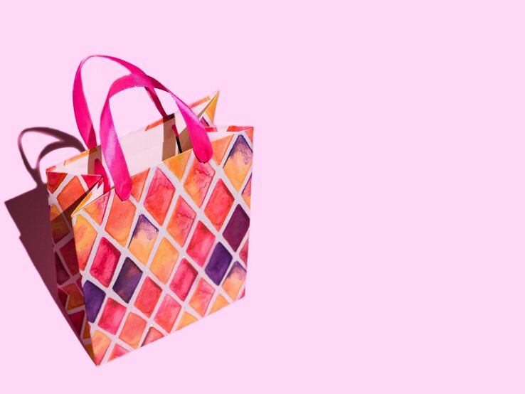 Bunte Geschenktüte mit pinken Griffen und rautenförmigem Muster in Rot-, Orange- und Lilatönen auf rosa Hintergrund.