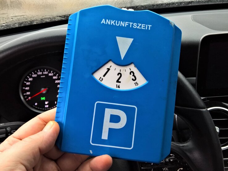 Eine Hand hält eine blaue Parkscheibe mit der Ankunftszeit auf 14 Uhr vor einem Autolenkrad und einem Tacho.