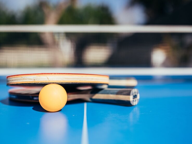 Zwei Tischtennisschläger und ein Ball liegen auf einem blauen Tisch vor unscharfem Hintergrund im Freien.