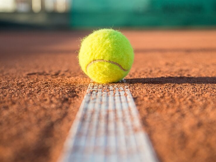 Ein gelber Tennisball liegt knapp über der weißen Linie auf einem sandigen Tennisplatz in der Sonne.