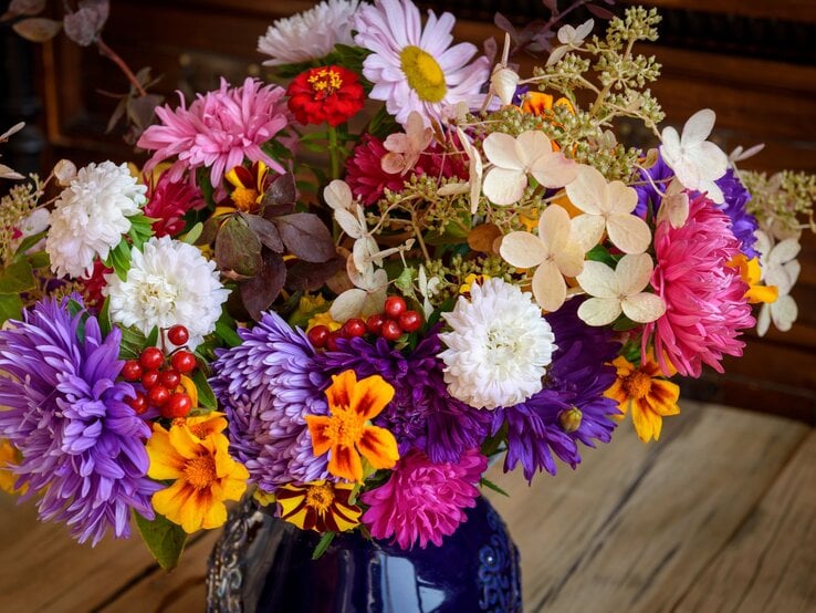 Ein bunter Blumenstrauß aus Chrysanthemen, Zinnien und Astern mit leuchtenden Herbstfarben in einer blauen Vase auf einem Holztisch.