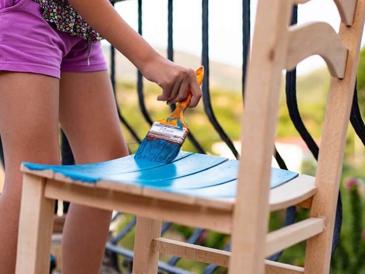 Eine Person in lila Shorts streicht mit einem blauen Pinsel eine hölzerne Stuhllehne auf einem sonnigen Balkon an.