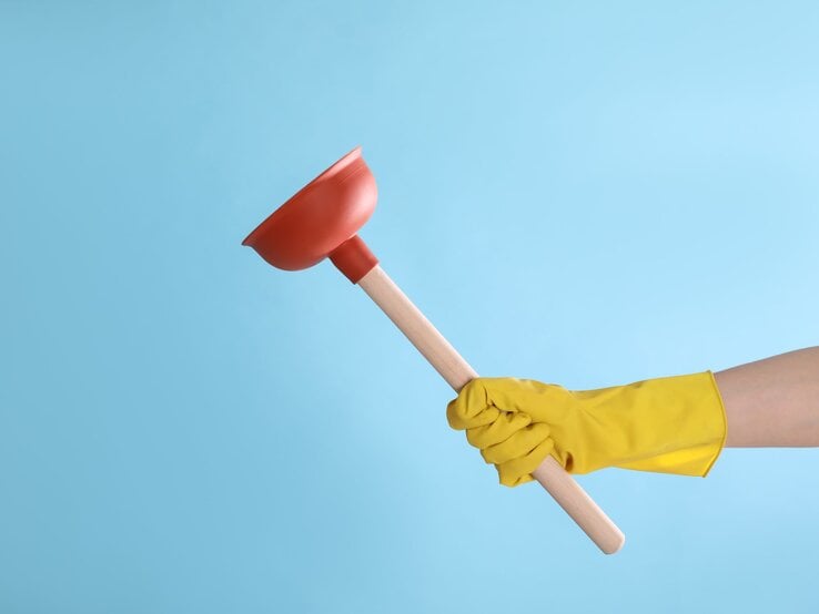 Eine Hand in gelbem Gummihandschuh hält einen roten Pümpel mit Holzgriff vor einem blauen Hintergrund.