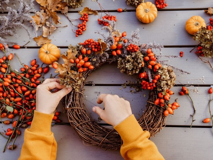 Hände in gelbem Pullover dekorieren einen herbstlichen Kranz aus Hagebutten, Trockenblumen und Eichenblättern auf Holztisch.