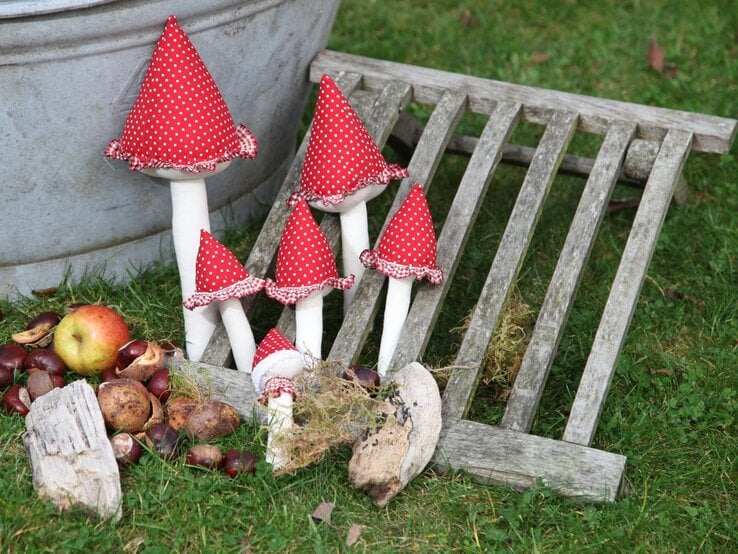 Fünf dekorative Stoffpilze mit roten, gepunkteten Hüten stehen neben Kastanien, Holzstücken und einem Apfel auf grünem Rasen.