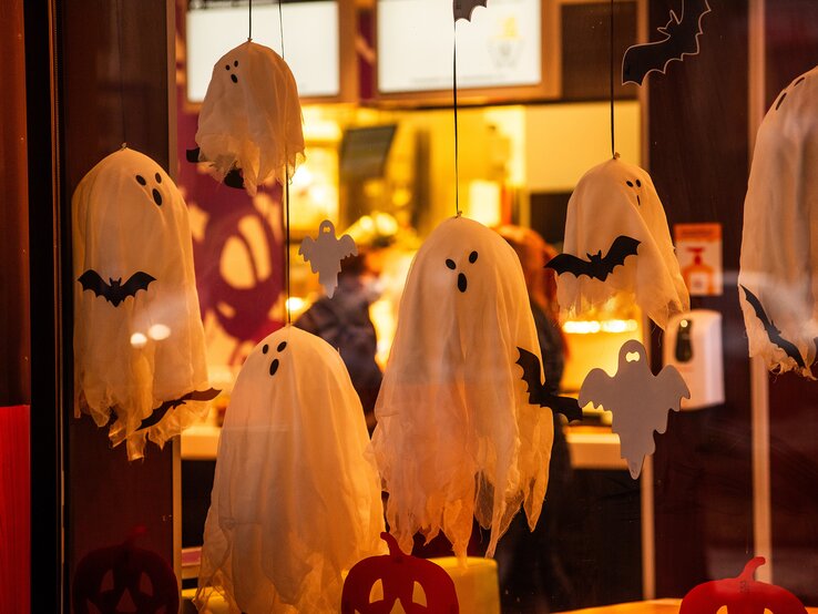 Weiße Geister und schwarze Fledermäuse aus Stoff hängen als Halloween-Dekoration hinter einer erleuchteten Glasscheibe.