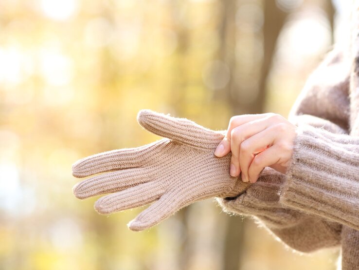 Eine Person zieht sich bei warmem Herbstlicht beige Strickhandschuhe über, im Hintergrund ein Wald in sanften Unschärfen.