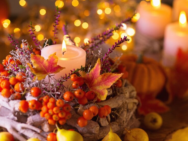 Ein brennendes Teelicht steht in einem herbstlichen Gesteck aus bunten Blättern, Beeren und Zweigen vor leuchtenden Lichterketten.