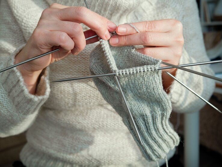 Nahaufnahme von Händen, die gerade dabei sind, eine Socke aus hellgrauer Wolle zu stricken. Die Person hält mehrere Stricknadeln.