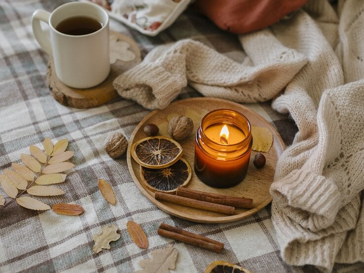 Eine entzündete kerze steht auf einem Tablet. Ein Kaffee daneben und eine Strickjacke. Alles ist in gemütliche braune Herbsttöne getaucht.