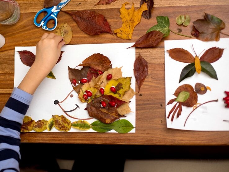 Ein Kind legt bunte Herbstblätter und rote Beeren auf ein Papier, um ein Igelbild zu gestalten, daneben eine Schere und Blätter.