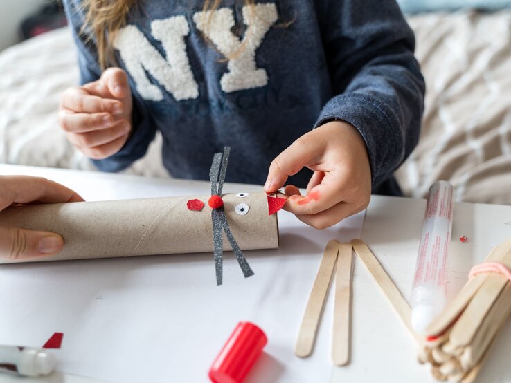 Ein Kind bastelt mit Küchenrolle, Kleber und bunten Materialien eine Figur auf einem weißen Tisch.