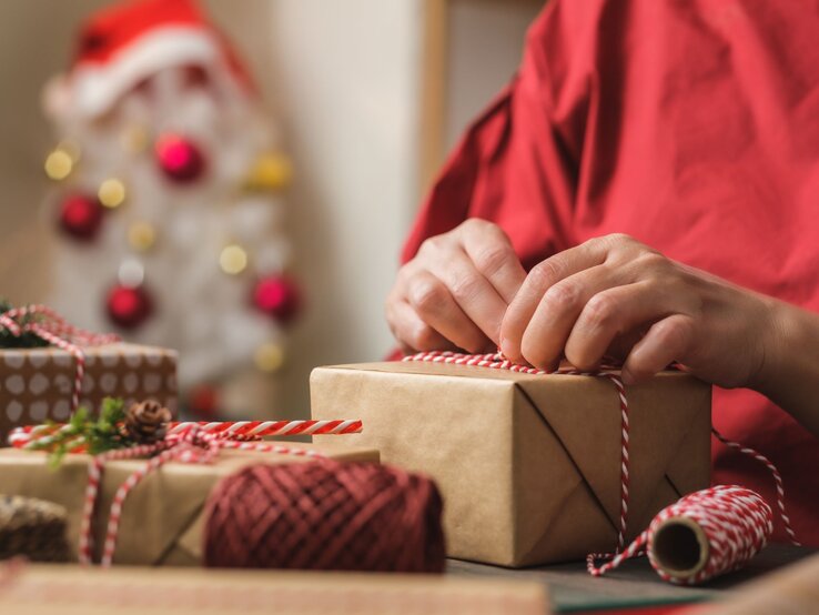 Eine Person bindet eine rot-weiße Schnur um ein Geschenk, im Hintergrund ein unscharfer Weihnachtsbaum mit Kugeln.