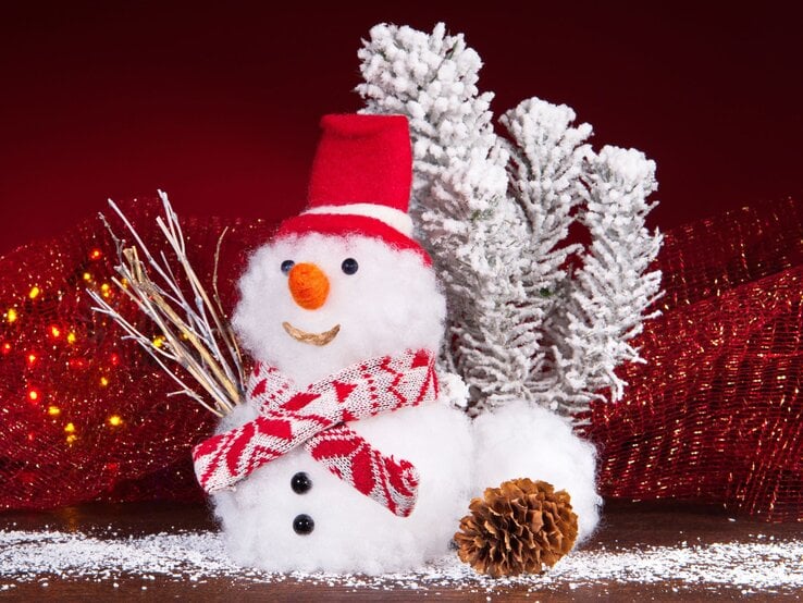 Ein lächelnder Schneemann mit rotem Hut und Schal steht vor schneebedeckten Zweigen, umgeben von roten Lichtern und einer Kiefernzapfe.