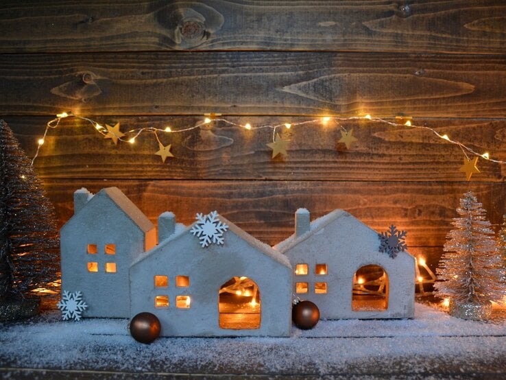 Kleine, weiße Häuser aus Ton mit Schneeflocken-Deko und Lichterkette stehen auf Kunstschnee vor einer Holzwand, umgeben von glitzernden Tannen.