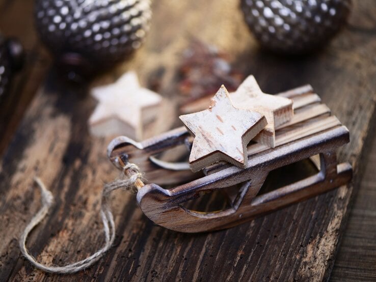 Kleine Holzschlitten-Dekoration mit zwei sternförmigen Anhängern auf rustikalem Holztisch, im Hintergrund unscharfe Weihnachtskugeln. Zwei weiße Holzsterne liegen auf einem kleinen Schlitten aus Holz, rustikaler Hintergrund mit Weihnachtsdeko. Detailaufnahme eines Mini-Holzschlittens mit Stern-Dekoration, rustikaler Holzuntergrund und verschwommene Ornamente im Hintergrund.