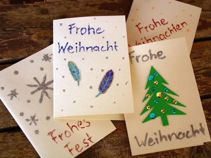 Handgemachte Weihnachtskarten mit Glitzersternen, Federdekoration und grünem Tannenbaum auf rustikalem Holztisch arrangiert.