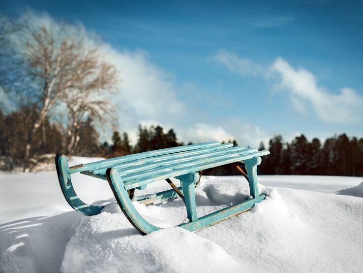 Ein türkisfarbener Holzschlitten steht verlassen auf frischem Schnee vor einem klaren, blauen Himmel und schneebedecktem Wald.