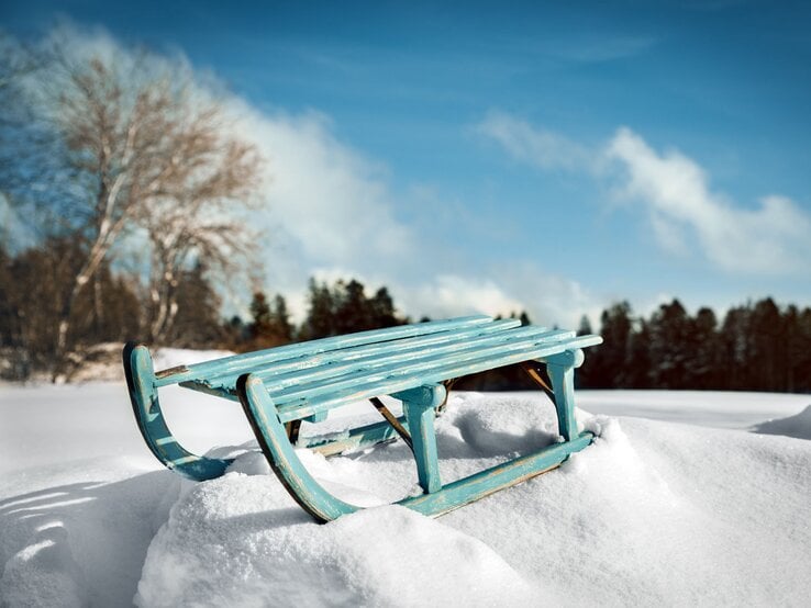 Ein türkisfarbener Holzschlitten steht verlassen auf frischem Schnee vor einem klaren, blauen Himmel und schneebedecktem Wald.