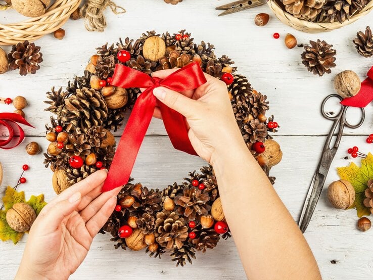 Zwei Hände binden eine rote Schleife an einen festlichen Adventskranz aus Tannenzapfen, Nüssen und roten Beeren auf hellem Holztisch.