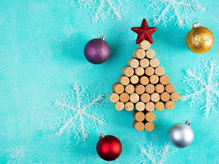 Aus Weinkorken gelegter Weihnachtsbaum mit rotem Stern, umgeben von Christbaumkugeln und weißen Schneeflocken auf türkisfarbenem Hintergrund.