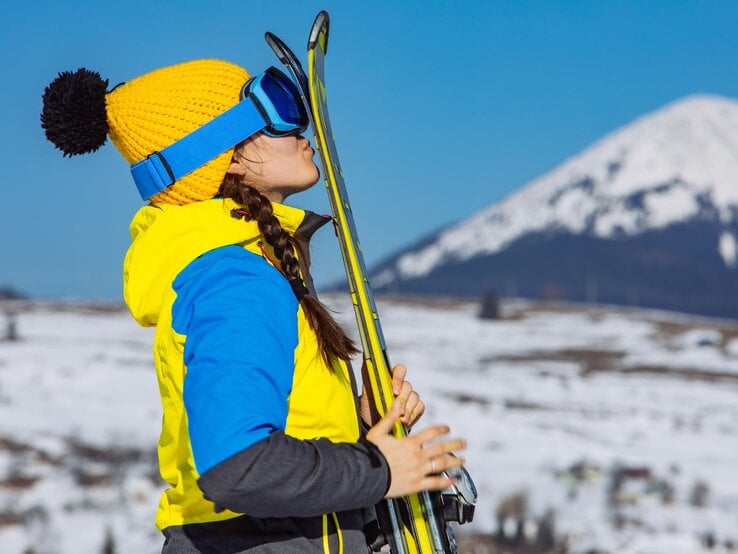 Junge Frau in gelb-blauer Jacke und gelber Mütze küsst im Schnee lächelnd ihre Skier, Berglandschaft im Hintergrund.