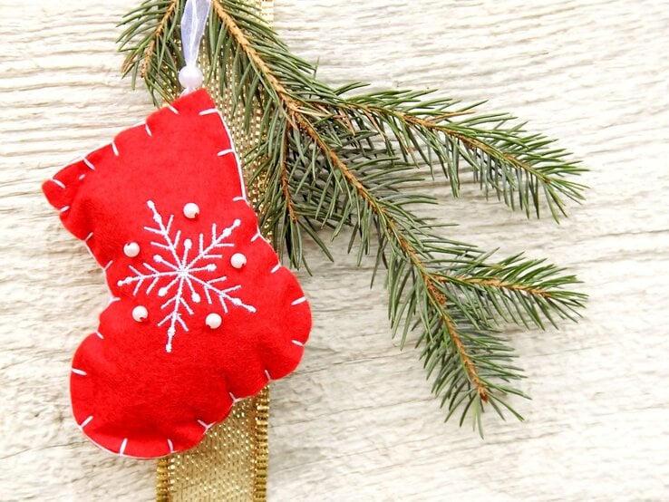 Eine rote Filz-Weihnachtssocke als Nikolausstiefel mit weißem Schneeflockenmotiv und Perlen hängt an einem Tannenzweig vor hellem Holz.