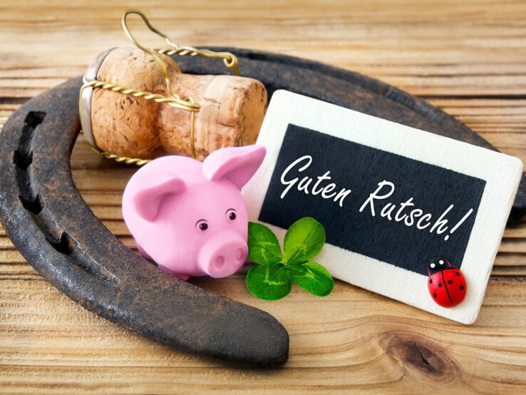 Ein rosafarbenes Glücksschwein, ein Kleeblatt, ein Hufeisen und eine Holztafel mit der Aufschrift „Guten Rutsch!“ liegen auf hellem Holz.