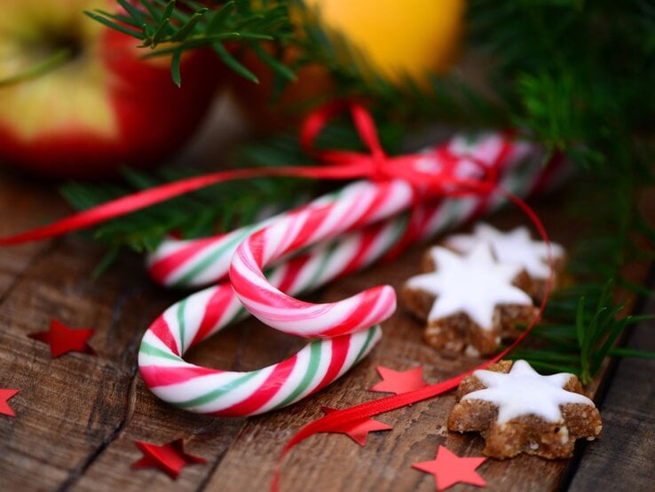 Zwei rot-weiß-grüne Zuckerstangen liegen auf Holz neben Zimtsternen, Sternkonfetti in Rot und grünen Tannenzweigen.