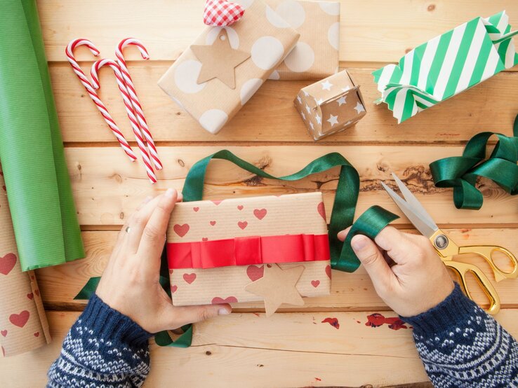 Zwei Hände in blauem Strickpulli verpacken ein Geschenk in Herzpapier mit rotem Band, umgeben von Geschenkpapier, Schleifen und Zuckerstangen.