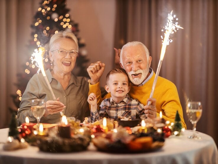 Älteres Paar und ein Junge sitzen an Silvester lächelnd mit Wunderkerzen an einem festlich gedeckten Tisch vor einem geschmückten Weihnachtsbaum.