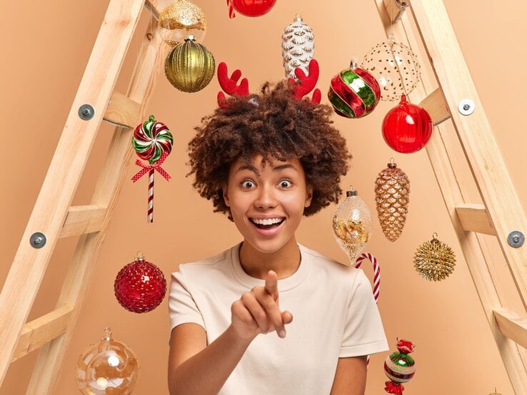 Junge Frau mit lockigem braunem Haar und Rentiergeweih lächelt überrascht, unter einer geschmückten Holzleiter, die als Alternative zum Weihnachtsbaum funktioniert. 