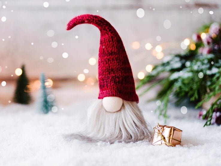 Ein selbst gebastelter Wichtel aus Socken mit roter Strickmütze und grauem Bart steht auf schneebedecktem Boden, daneben ein goldenes Geschenk.