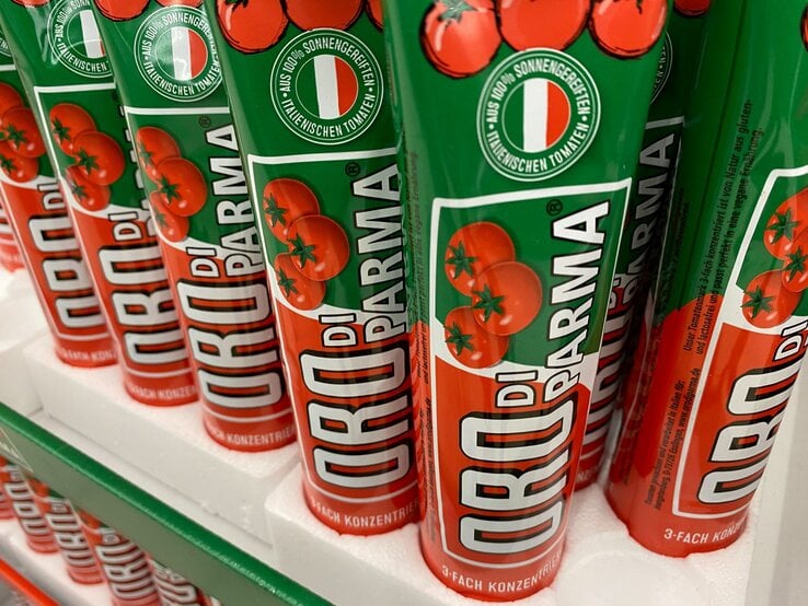 Mehrere rote Tuben Oro di Parma Tomatenmark mit italienischem Logo und Tomatenabbildung stehen auf weißem Karton in einem Regal.