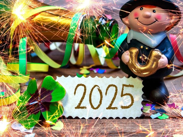 Glücksbringer-Figur mit goldenem Hufeisen, Kleeblatt, bunten Luftschlangen und Wunderkerzen vor einer Karte mit „2025“.