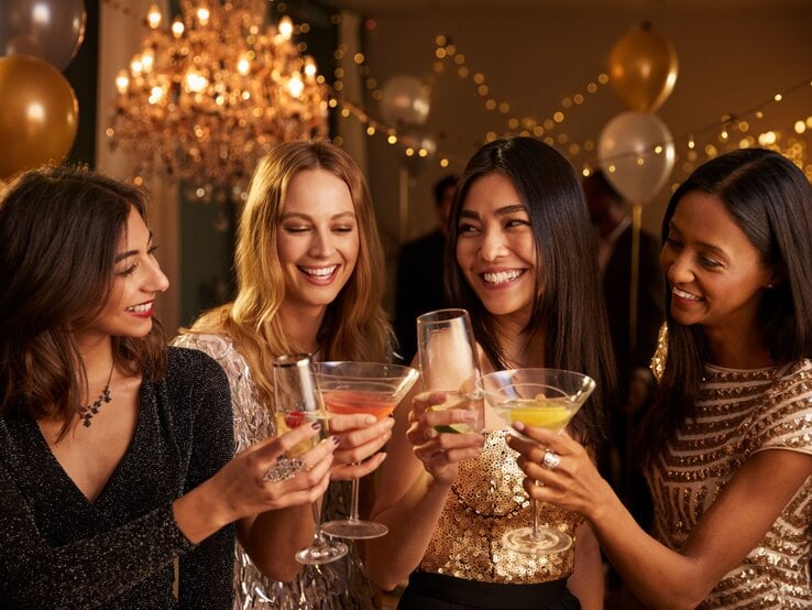 Vier junge Frauen in glitzernden Abendkleidern stoßen lachend mit Cocktails in einem festlich beleuchteten Raum an.