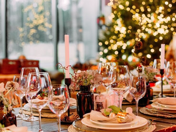 Festlich gedeckter Tisch mit eleganter Weihnachtsdeko aus goldenen Serviettenringen, Kerzen und Tannenzapfen vor leuchtendem Weihnachtsbaum.