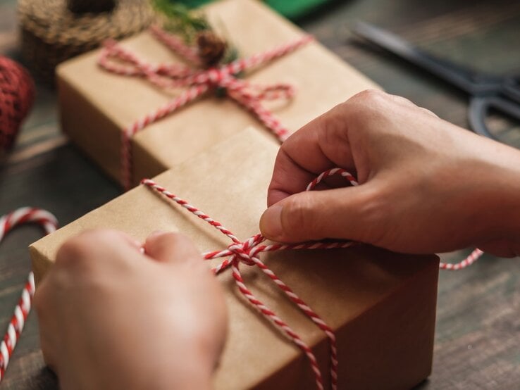 Nahaufnahme von Händen, die ein in Packpapier verpacktes DIY-Weihnachtsgeschenk sorgfältig mit rot-weißer Schnur verzieren.