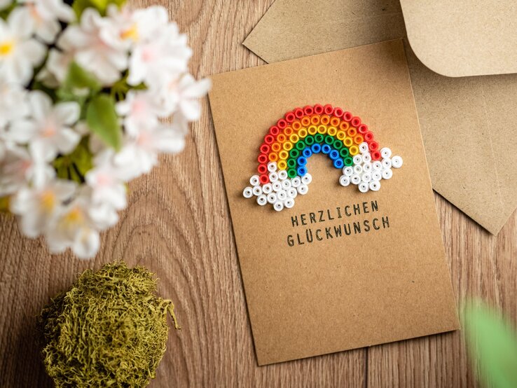 Glückwunschkarte aus braunem Papier mit Regenbogen aus bunten Bügelperlen, umgeben von Blumen und Moos auf Holztisch.