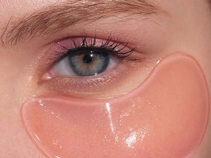 Ein blau-graues Auge mit langen Wimpern, zartem Lidschatten und einer glänzenden rosa DIY-Augenpad unter dem unteren Lid.