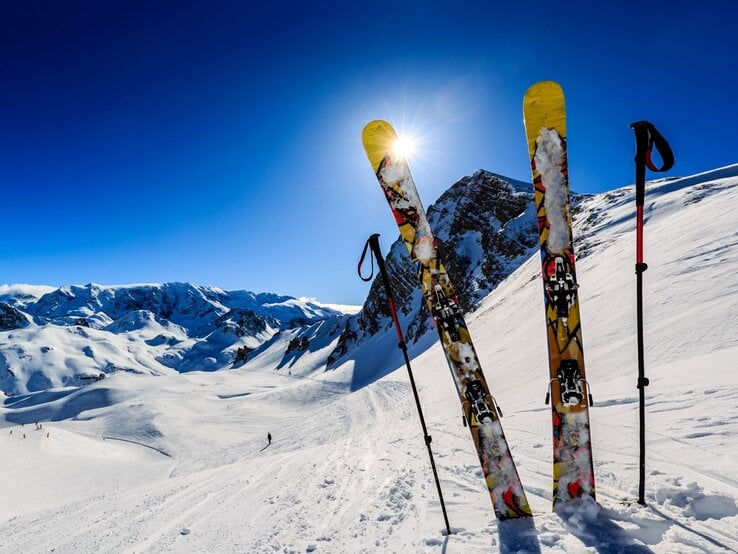 Zwei gelbe Ski und zwei Stöcke stehen im glitzernden Schnee vor einem sonnigen Bergpanorama mit tiefblauem Himmel.