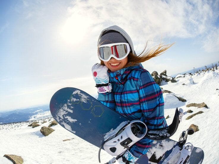 Eine junge Frau mit weißer Skibrille und kariertem blauen Anorak lächelt auf einer verschneiten Berglandschaft mit Snowboard in der Hand.