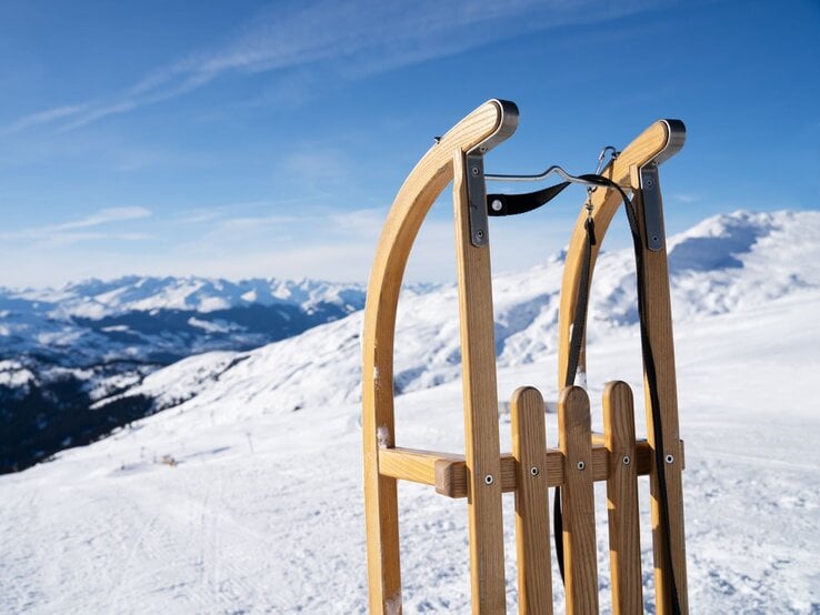 Ein hölzerner Schlitten steht auf schneebedecktem Bergplateau, im Hintergrund erstrecken sich sonnige Alpen unter klarem Himmel.