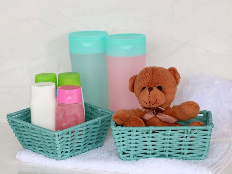 Stoffteddy in einem Korb, daneben Baby-Pflegeprodukte in Plastikflaschen und ein weißes Netzschwämmchen auf einem Handtuch.