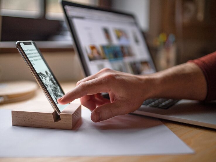 Eine Hand in rotem Ärmel berührt ein Smartphone, das auf einer DIY-Handyhalterung steht, im Hintergrund ein Laptop auf einem Schreibtisch.