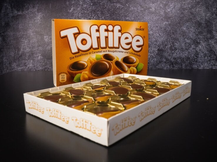 Geöffnete Packung Toffifee mit goldenen Förmchen und Schokoladenpralinen, dahinter die braune Verpackung auf schwarzem Untergrund.