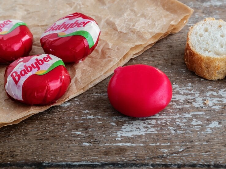 Nahaufnahme von Babybel-Käse auf rustikalem Holztisch mit Pergamentpapier und einer Baguettescheibe im Hintergrund.