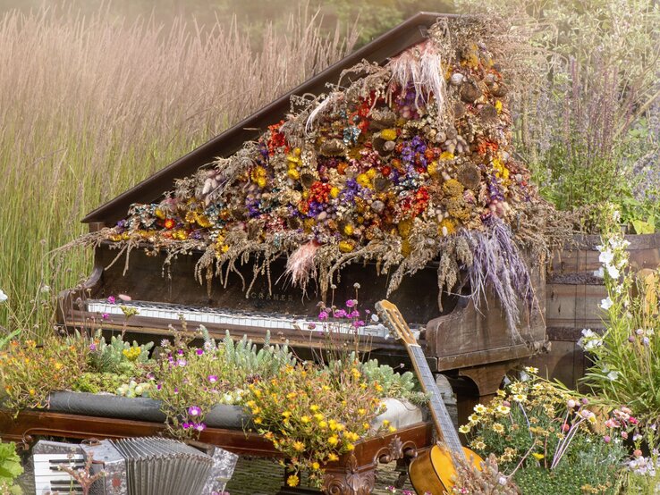 Ein antikes Klavier, überwuchert mit bunten Trockenblumen und Gräsern, steht inmitten einer idyllischen Wiese mit blühenden Wildblumen.